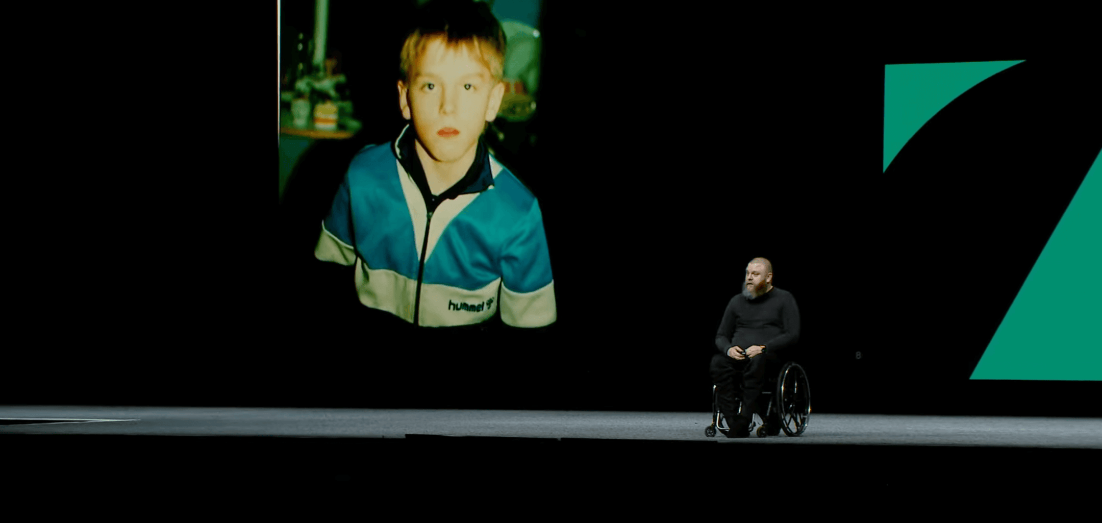 Halli, dressed in black sitting in a wheelchair, on stage at Config. Behind him on a big screen is a photo of himself as a child.
